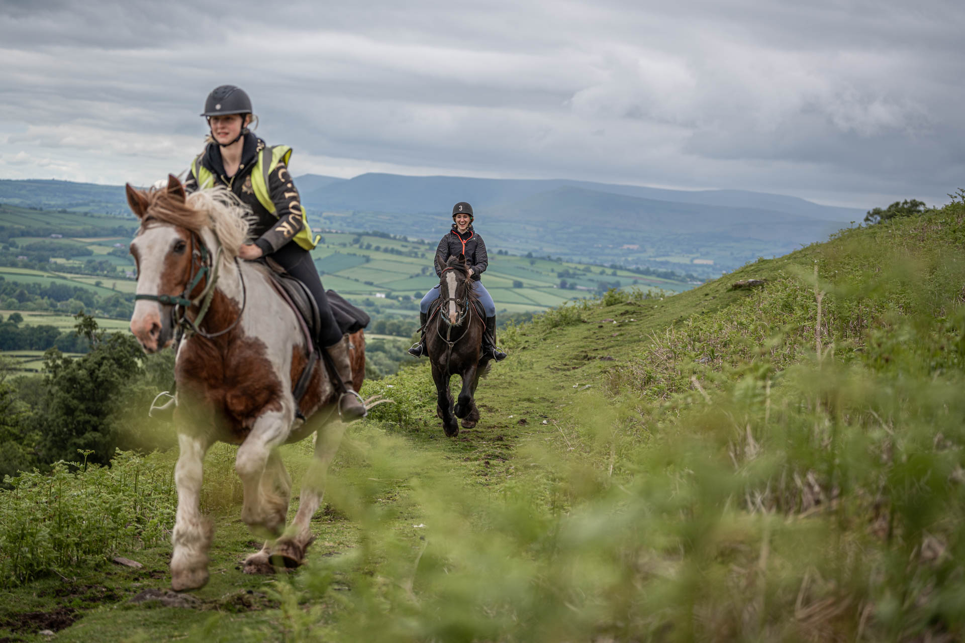 An image of two riders galloping towards the camera over the Brecon Beacons, with an expansive view behind.