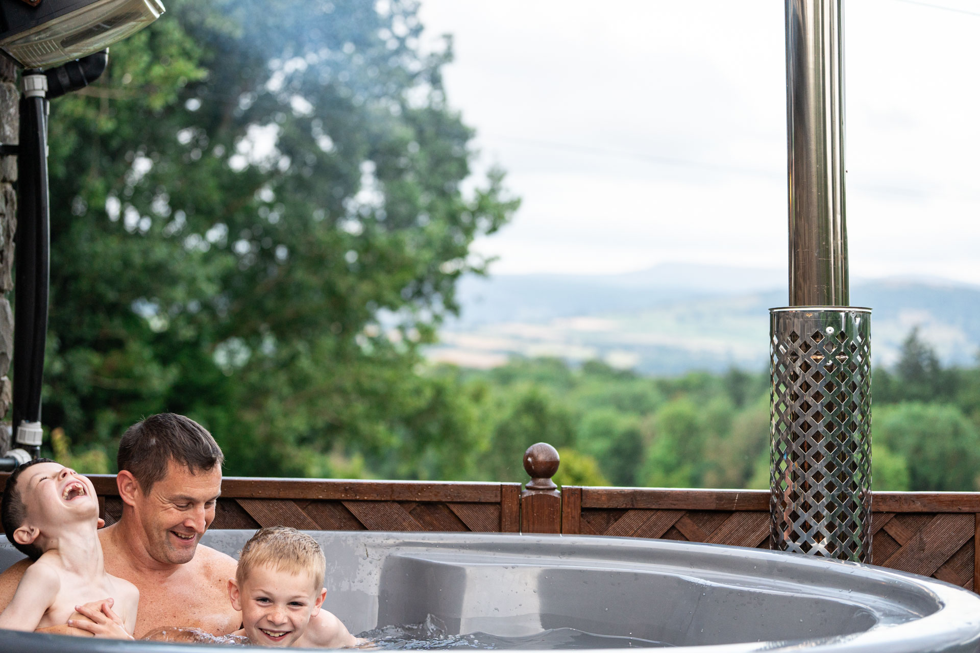 An image of a dad laughing with his two sons in a woodfired hot tub, with luscious greenery behind them.