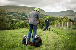 An image of two men on a Segways going down a mown grass path with a beautiful view of the Brecon Beacons in the background.