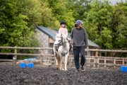 An image of a girl riding a white pony around Cantref Riding Centre's training area, the pony is being led round by the girl's mum.