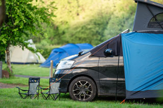 A close up image of a campervan and camping chairs on a grass pitch at Cantref with greenery surrounding them.