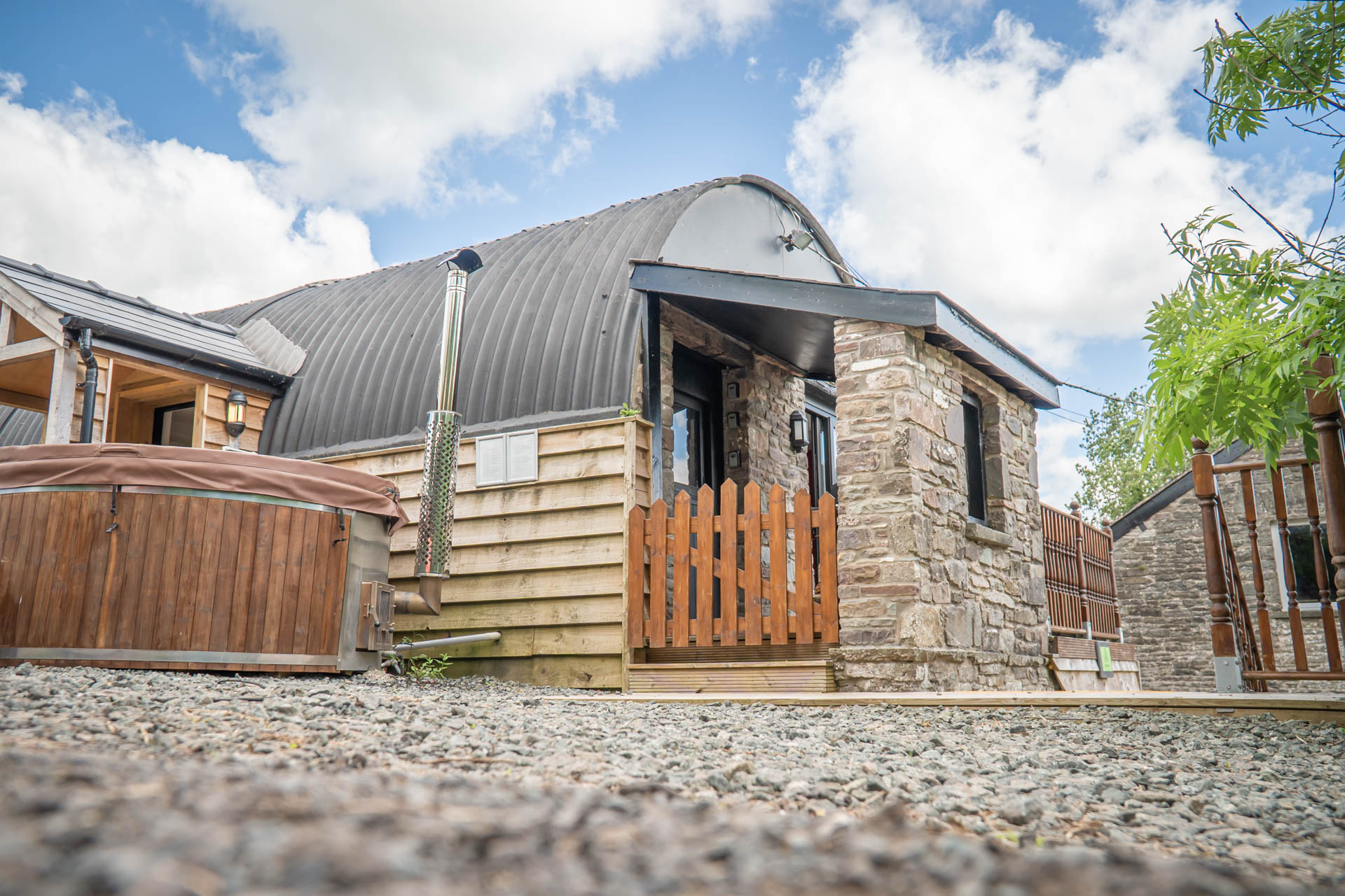 An image of a hayloft with a tin Barrell Vault roof and exterior wooden cladding. Converted into accommodation with an entrance porch and outdoor wooden fired hot tub.
