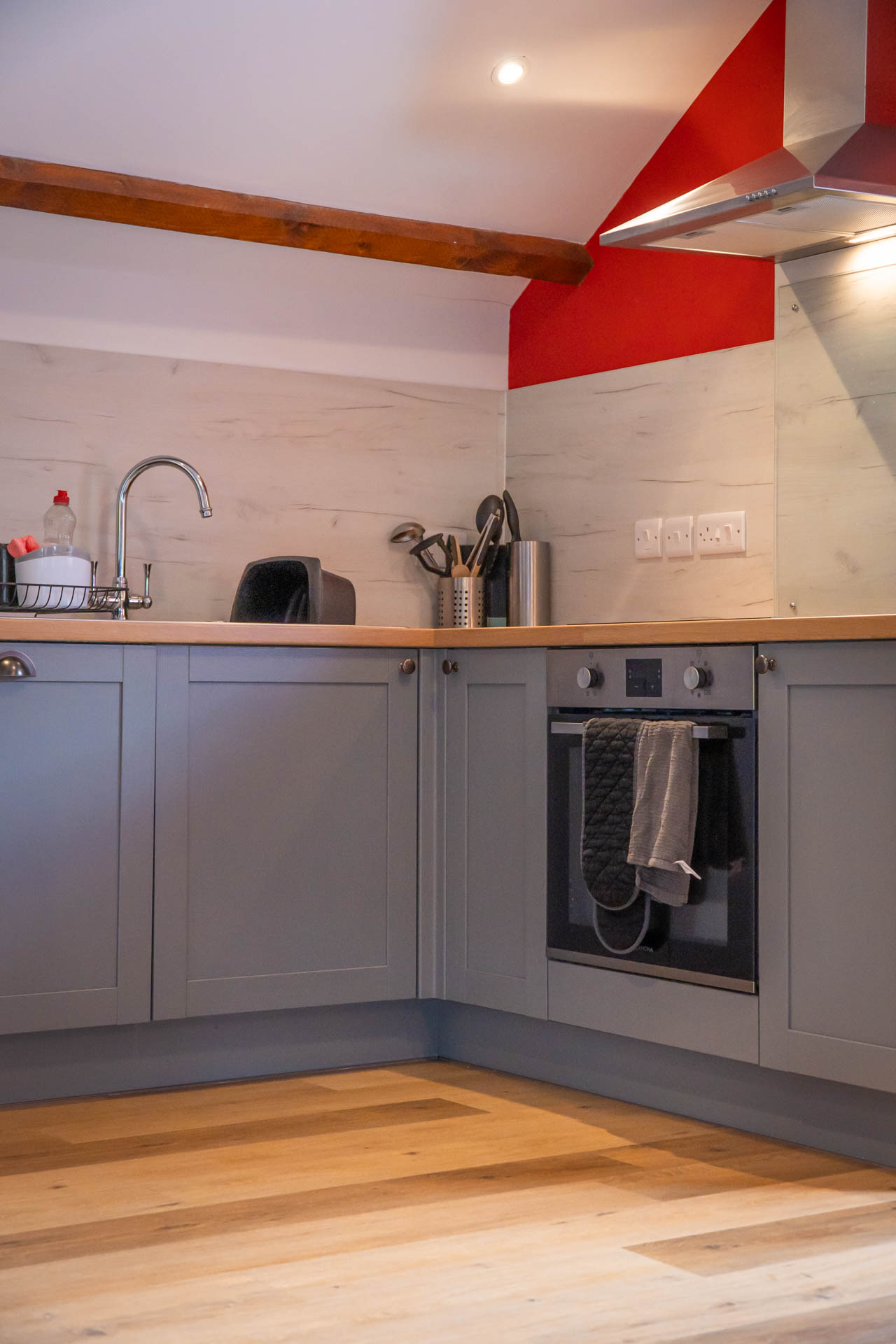 An image of a grey kitchen with wooden worktops and white splashback, set into the pitched roof of the old Milking Barn with wooden beams exposed.