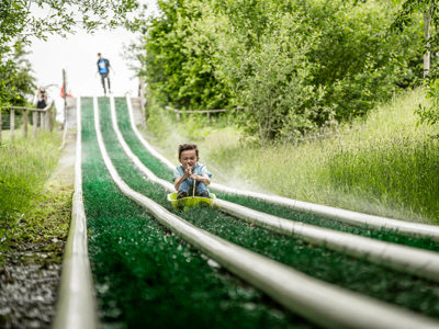 An action shot of a boy zooming down a wet artificial grass slide in a sledge, whilst being sprayed in the face with sprinklers.