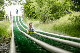 An action shot of a boy zooming down a wet artificial grass slide in a sledge, whilst being sprayed in the face with sprinklers.