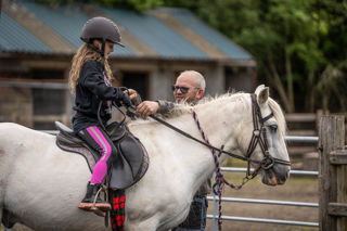 An image of a girl seated on a small white horse, having the reins checked by a member of the Cantref team.
