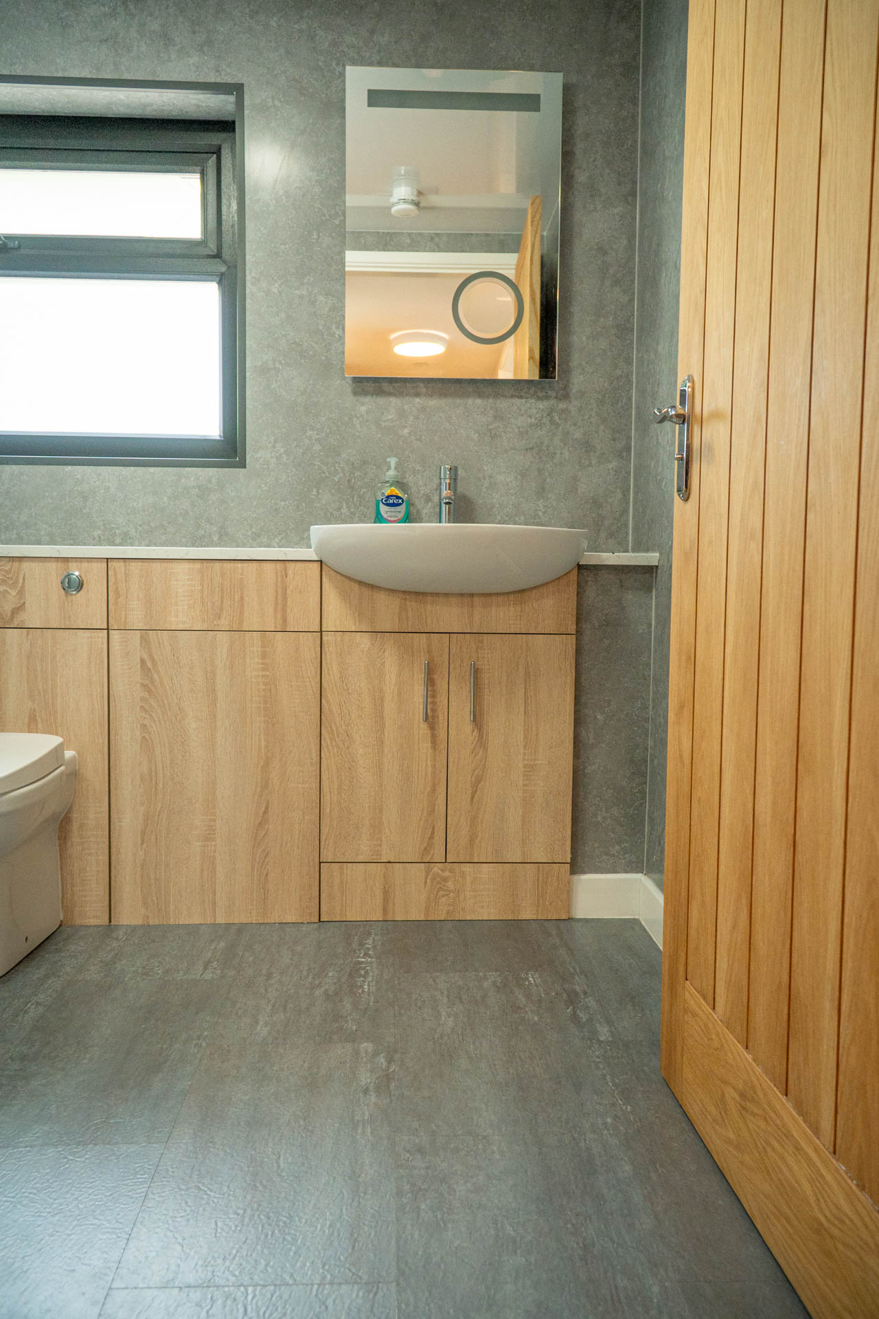 An image of a light, spacious, family bathroom with stone effect walls, grey wood effect flooring, frosted window, sink and wc.
