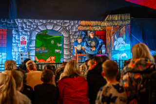 An image of a seated crowd gathered in front of a stage, with a woman delivering a show about bottle feeding lambs.