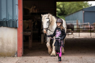 An image of a girl wearing a riding helmet, leading a small white horse out of the stable.
