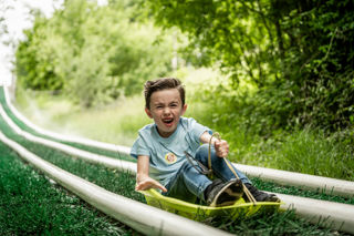 An action shot of a boy zooming down a wet artificial grass slide in a sledge.