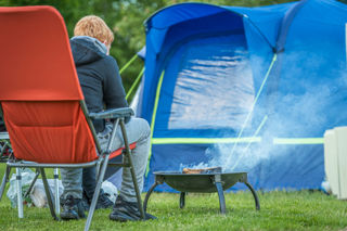 An image of a camper sat on a red camping chair tending to an elevated fire pit, with their blue tent in the background.