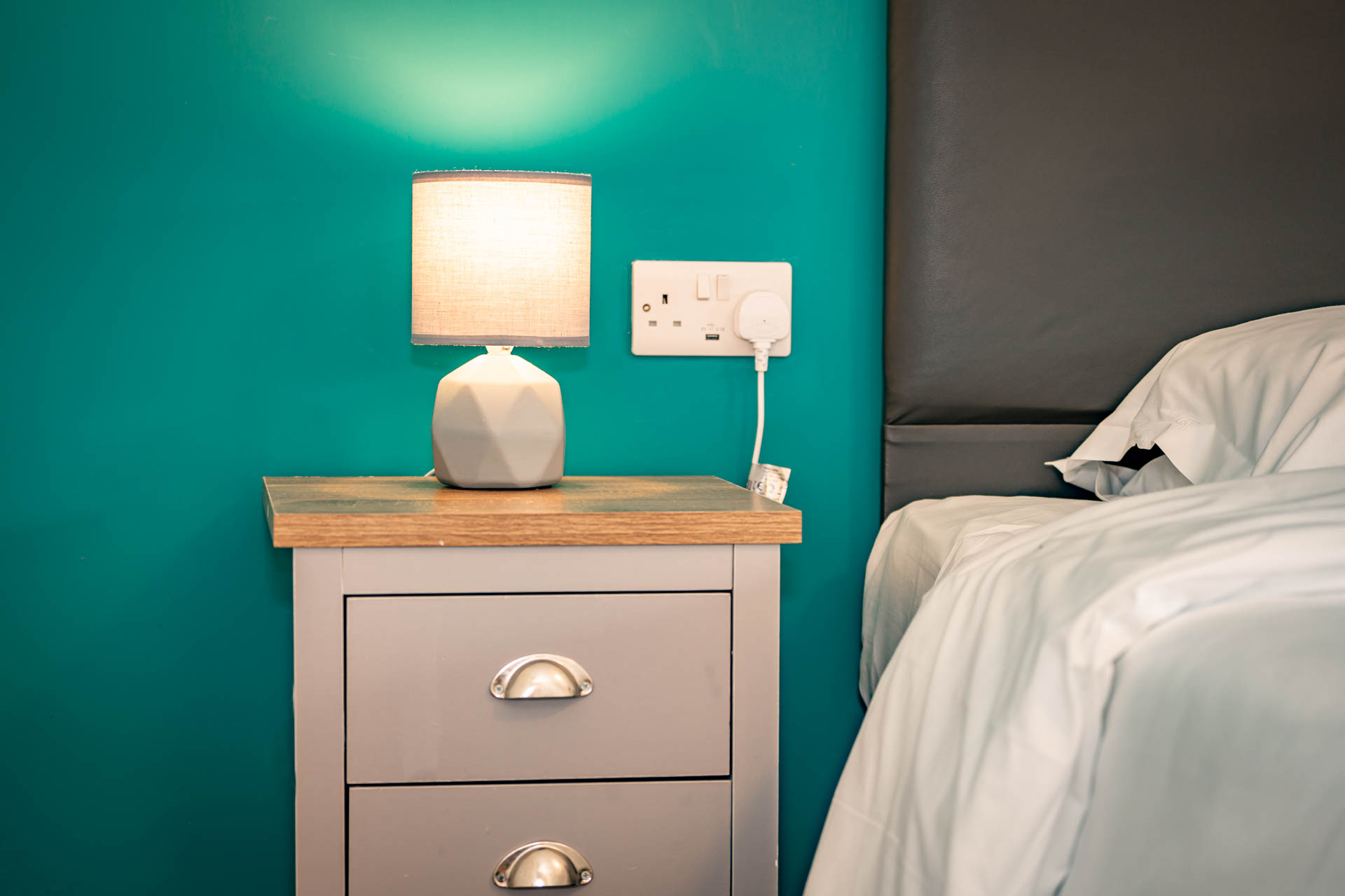 A close up image of a bedside table, lamp and bed.