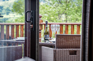 An image looking through open French doors onto a veranda, with stunning views. There's a set dining table, a bottle of bubbly and some glasses.