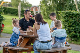 An image of a family of five enjoying their food from a picnic bench, surrounded by gorgeous views and outdoor play.