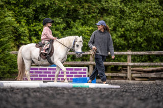 An image of a girl riding a white pony around Cantref Riding Centre's training area, the pony is being led round by the girl's mum.