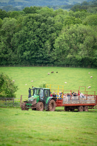 An image of a green John Deere tractor pulling a red and yellow trailer filled with people. They're enjoying a ride over the farm land at Cantref Adventure Farm, with beautiful rolling views in the distance and sheep grazing.