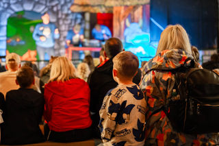 An image of a seated crowd gathered in front of a stage, with a woman delivering a show about bottle feeding lambs.