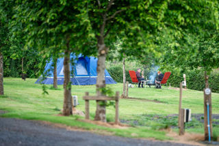 An image of two campers sat on camping chairs, tending to an elevated fire pit, getting ready to barbeque some food, surrounded by greenery.