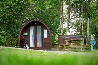 An image of an arched dark wooden pod, surrounded by trees and foliage. Adjacent there's a woodfired hot tub, barbeque and picnic bench.