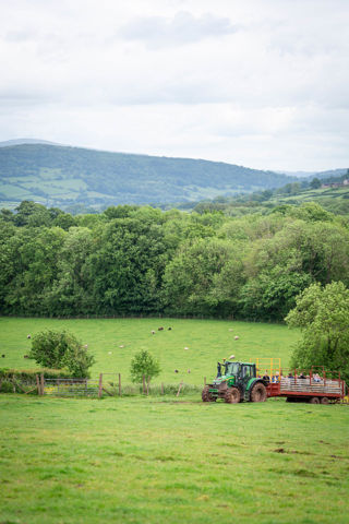 An image of a green John Deere tractor pulling a red and yellow trailer filled with people. They're enjoying a ride over the farm land at Cantref Adventure Farm, with beautiful rolling views in the distance.