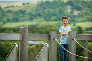 A close up image of a boy standing on top of a wooden platform, with a big smile on his face and a stunning panoramic view of the Brecon Beacons behind him.