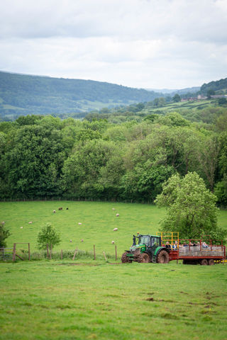 An image of a green John Deere tractor pulling a red and yellow trailer filled with people. They're enjoying a ride over the farm land at Cantref Adventure Farm, with beautiful rolling views in the distance.