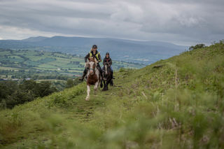 An image of two riders galloping towards the camera over the Brecon Beacons, with an expansive view behind.