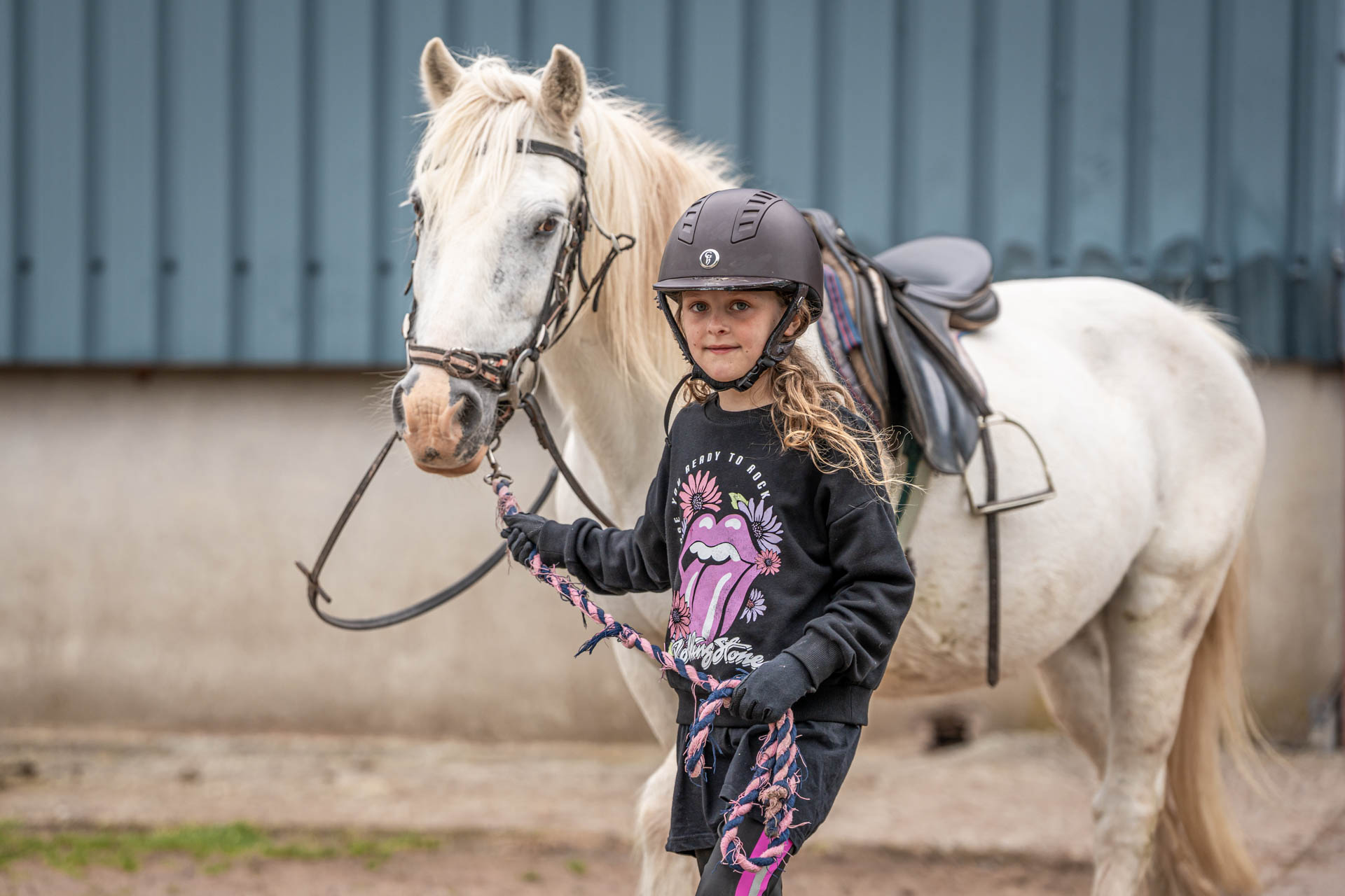 An image of a girl wearing a riding helmet, leading a small white horse out of the stable.