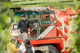 An image of a child sitting in the cab of a red combine harvester, pretending to drive it, whilst family watch on from outside.