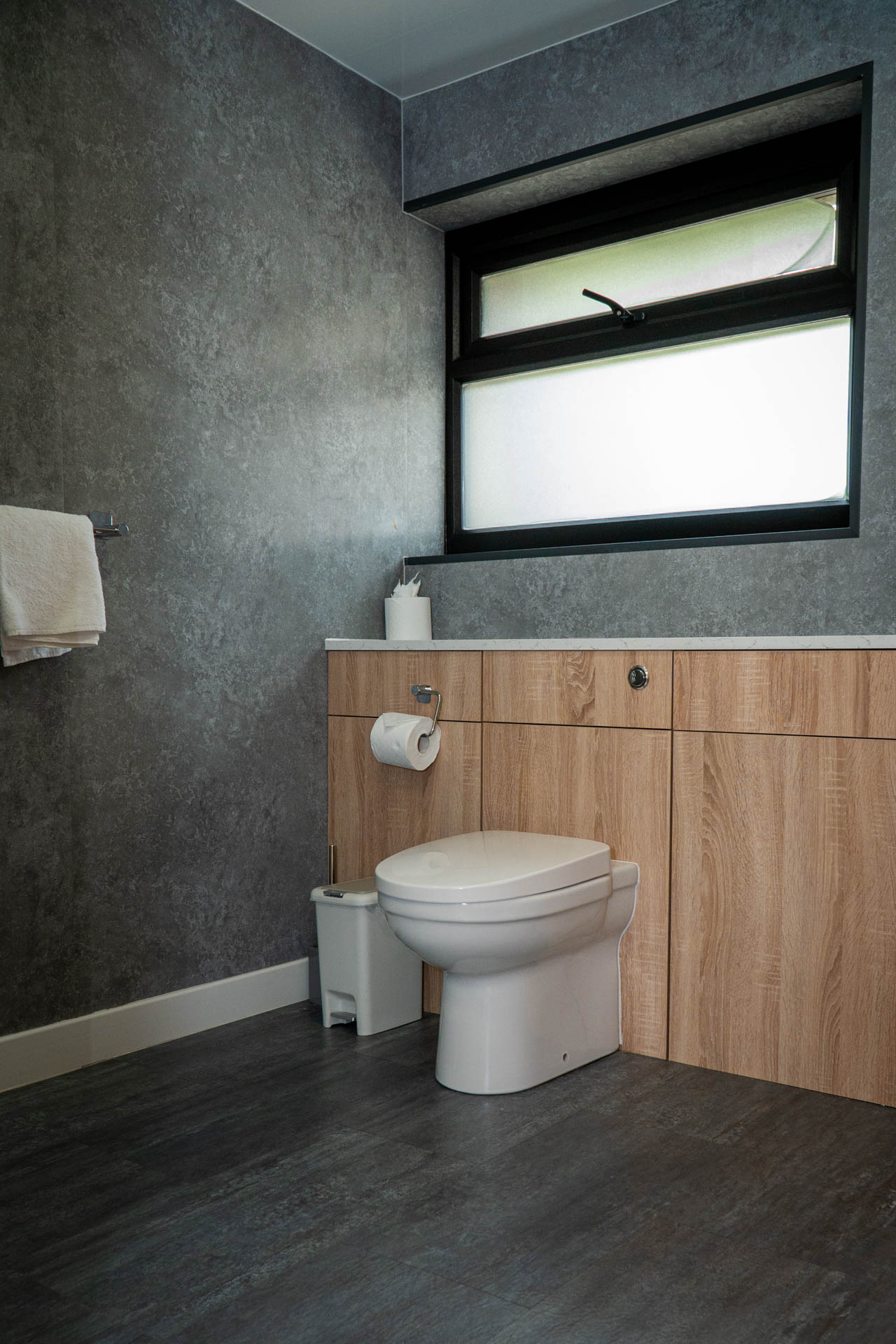 An image of a spacious family bathroom with frosted window, grey stone effect walls and dark grey wood effect flooring.